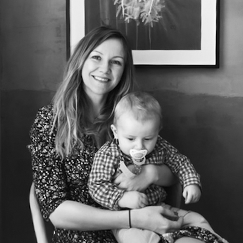 This is a photograph showing Claudia Petzinger smiling and holding a baby with a pacifier on her lap.
